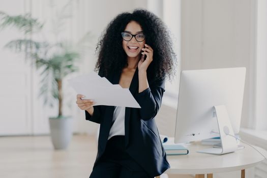 Intelligent woman with Afro hairstyle, holds paper documents, checks information, calls business partner via cell phone, analyzes future strategy, speaks with banking manager, dressed in black suit