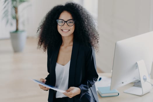 Photo of lovely African American woman holds some papers, being consultant manager, wears elegant clothes and spectacles, stands near desktop with computer and textbooks. Happy businesswoman