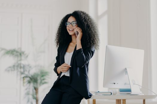 Happy female entrepreneur shares ideas for project with colleague, holds modern cell phone, makes phone call, wears spectacles and formal suit, poses in office, looks somewhere. Technology, business