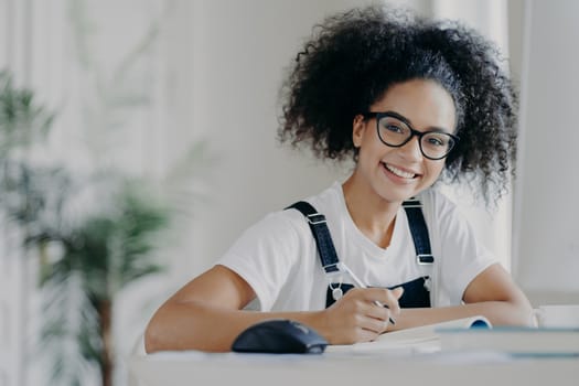 Time for studying. Happy dark skinned teenage girl holds pen, writes down information needed for course work, busy preparing for classes, dressed in casual wear and spectacles, being hard working