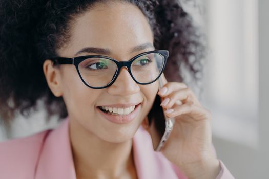 Headshot of beautiful smiling dark skinned woman wears optical glasses, holds modern cell phone, has well cared complexion, talks about future investments with business partner, looks aside.
