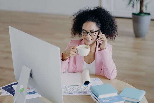 Glad dark skinned business lady looks happily at computer, drinks fresh hot beverage, holds modern mobile phone, dressed elegantly, poses against office interior, sits at desk with textbook, notepad