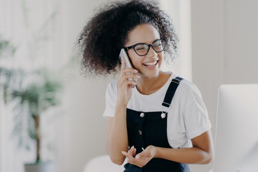 Photo of overjoyed female discusses something pleasant via cell phone, holds cellular near ear, being in good mood, wears glasses and casual clothing, poses in spacious room, blurred background