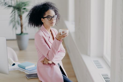 Indoor shot of thoughtful curly haired young woman holds cup of coffee, has pensive look at window, poses in coworking space, leans at table, being in office, dressed elegantly, wears spectacles