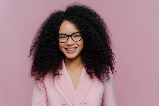 Close up portrait of happy curly haired woman has gentle smile on face, dark healthy skin bushy hair, wears elegant clothes, isolated on purple background, enjoys coming weekend, works in office