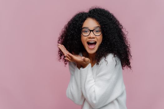 Overjoyed happy curly young woman with cheerful expression, keeps mouth widely opened, wears casual white jumper and optical glasses, poses over violet background. Emotions and happiness concept