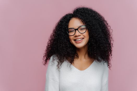 Headshot of pleased lovely woman with curly hairstyle, smiles gently at camera, wears optical glasses and white casual sweater, poses against purple background. Positive emotions and feelings concept