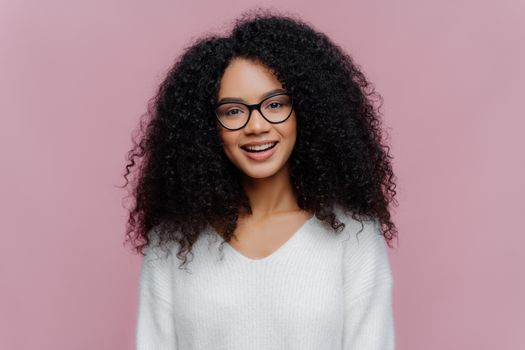Portrait of pleasant looking smiling woman with Afro hairstyle, wears optical glasses and white sweater, has cheerful face expression, isolated over purple background. Millennial girl poses indoor