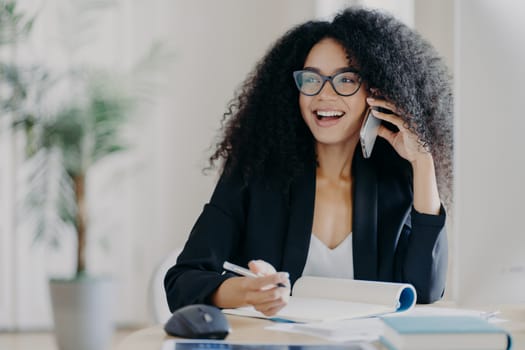 Photo of glad smiling dark skinned woman calls business partner, being in good mood, makes notes, sits at workplace, wears optical spectacles, formal suit, poses in cabinet. Work, business, technology