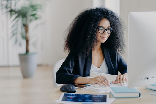 Freelance Afro woman works remotely, writes information, focused at computer screen with delighted expression, poses at workplace in spacious cabinet, potted plant in background. E learning concept