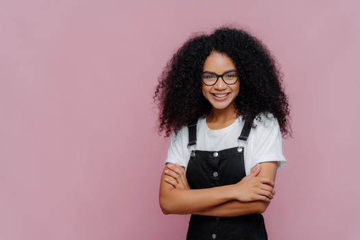 Photo of lovely teenage girl with Afro haircut, keeps arms folded, wears glasses, white t shirt and black overalls, looks gladfully at camera, stands against purple background with copy space for text