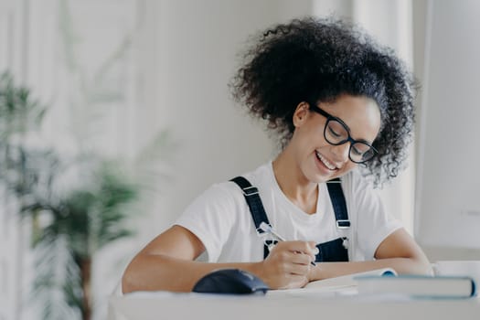 Happy Afro American female student writes down information, prepares for exams, sits in coworking space, has curly dark hair, wears optical glasses white t shirt and overalls, studies in spacious room