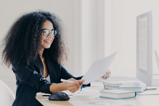 Glad Afro American woman with curly hairstyle, makes financial report, holds paper documents, has happy smile wears spectacles for vision correction dressed in elegant clothes computer screen in front