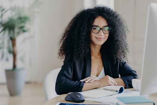 Positive female director with Afro curly hair, rewrites information in notepad from computer, wears optical glasses and formal wear, drinks coffee, poses at desktop, prepares working schedule