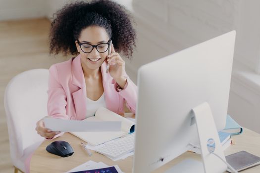 Pleased beautiful gorgeous curly haired businesswoman looks through documents, has telephone talk, sits at worplace, uses computer for making financial report, wears spectacles for vision correction