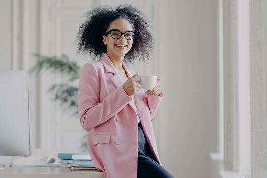 Photo of relaxed businesswoman holds cup of hot drink, has coffee break, stands near her workplace in spacious white cabinet wears spectacles long pink jacket works in office. Time for rest after work