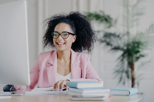 Horizontal shot of cheerful African American freelancer browses internet on computer, focused at screen, wears optical glasses and rosy formal costume, poses at desktop with pile of books, papers