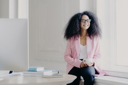 Happy female entrepreneur has bushy Afro hair, wears rosy jacket and black trousers, holds cup of drink, poses at windowsill in spacious cabinet with table, modern computer, enjoys aromatic coffee