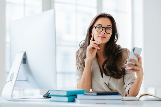 Female company office worker in formal wear uses modern gadget for work, poses at workplace near monitor, plans working schedule, browses information online, poses against window. Working time