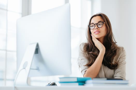 Indoor shot of serious brunette lady focused in monitor of computer, uses wireless internet for publication, keeps hand on cheek, has attentive look, sits at desktop, works on business contract