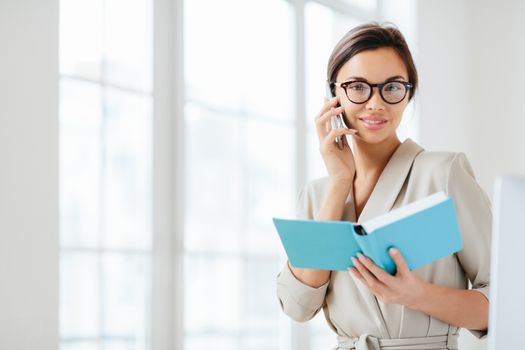 Photo of attractive woman with dark hair, has telephone conversation, agrees to meet with colleague, holds opened diary, wears glasses and formal suit, poses over white background plans daily schedule