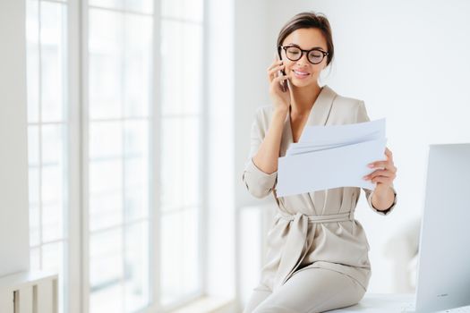 Attractive businesswoman reads papers or business documents, has telephone talk with business partner, examines paperwork before meeting, sits at desktop, smiles happily, works with documentation