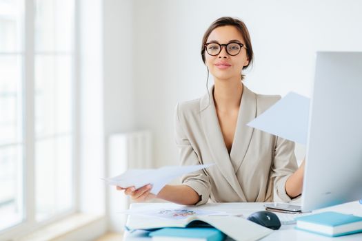 Busy successful female manager works with papers in office, poses at desktop, wears spectacles and formal outfit, busy preparing report, looks confidently at camera. Businesswoman reviews documents