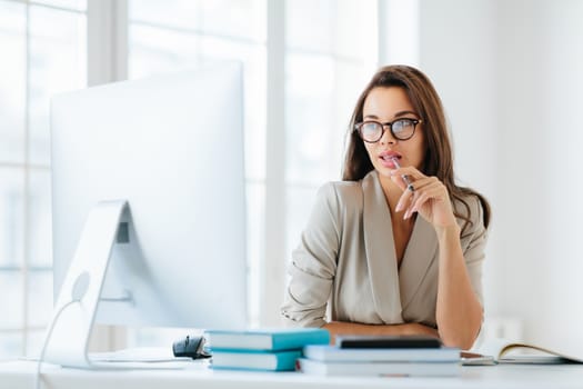 Contemplative female entrepreneur keeps pen in mouth, focused in monitor of computer, thinks on development of new strategy, wears elegant clothes and spectacles, poses against office interior