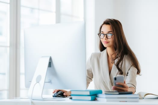 Pretty female entrepreneur with long dark hair, works on computer, develops strategy of advertising campaign, poses in modern coworking office, uses cellphone for chatting online, wears glasses