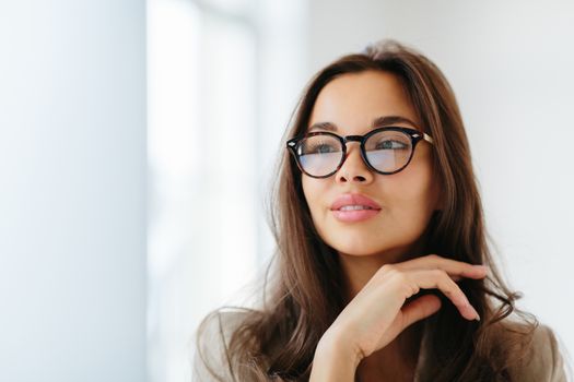 Close up shot of delicate woman wears optical glasses with dark frames, touches gently chin, looks aside thoughtfully, has dark straight hair, copy space on left side. Thoughtful businesswoman