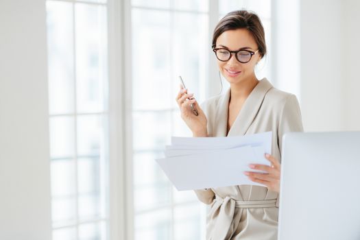 Photo of satisfied young woman with dark hair dressed in business suit, focused in papers, works in office, holds modern cellphone, wears optical glasses for good vision, has pleased expression