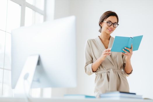 Cheerful female director holds personal organizer, looks through plans for day, wears beige formal costume, spectacles poses near dektop with big monitor white background verifies recorded information