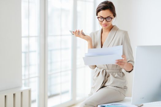 Photo of beautiful female entrepreneur in formal suit, focused in smartphone, checks information from paper documents, holds phone, going to search financial news online. Lawyer prepares for briefing