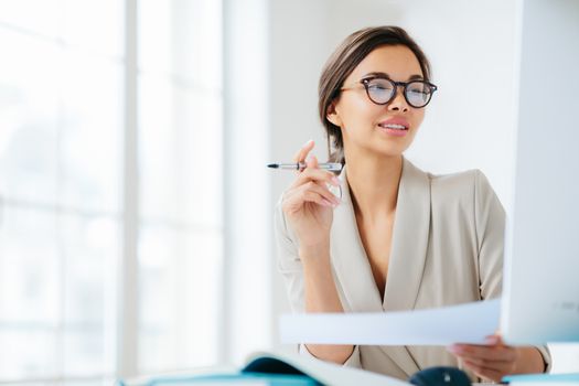 Prosperous businesswoman focused in monitor of computer, studies financial issues, holds document and pen, works in office, wears formal clothing and spectacles, poses over white background.