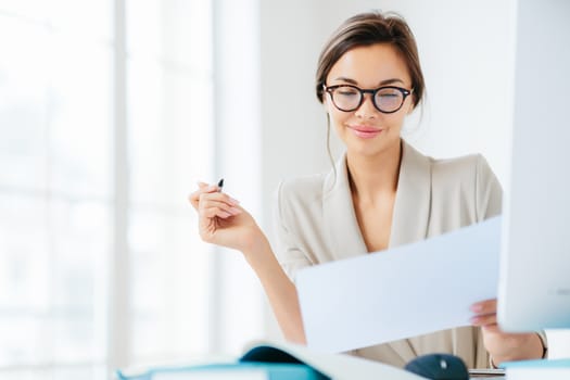 Concentrated successful businesswoman looks attentively in paper, studies terms of contract, holds pen, writes in documentations, dressed formally, poses at desktop against white spacious interior