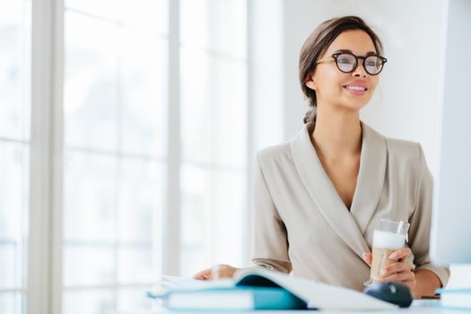 Positive woman works in office, sits at desktop, concentrated into monitor, drinks fresh milkshake, smiles and enjoys working, poses against office interior thinks about organization of business event