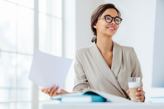 Cheerful businesswoman reads paper documents, focused aside with smile drinks milkshake sits at desktop makes business plan for project. Female executive manager in spectacles poses in coworking space