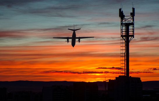 Silhouette of a low-flying plane over the city during sunset. The plane flies off into the sunset.