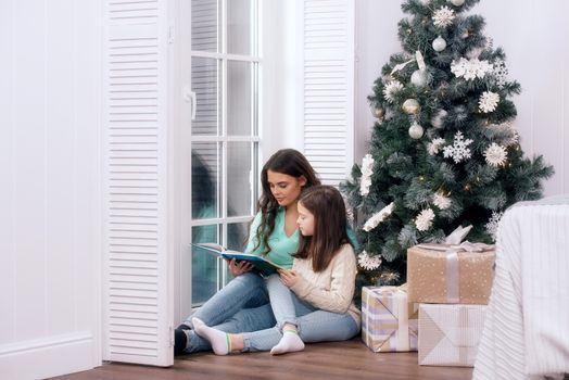 Mother and daughter reading a book sitting on the floor near christmas tree