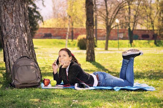 Beautiful young female student girl lying on the grass in a park, taking a break from studying and talking on the phone.