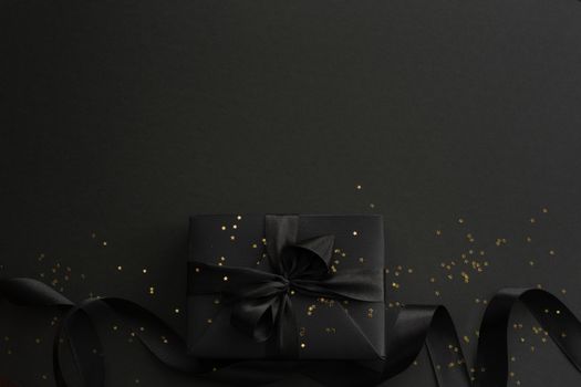 Black friday gift, paper box with silk ribbon bow and golden glitter stars on black paper background with copy space for text, flat lay top view template