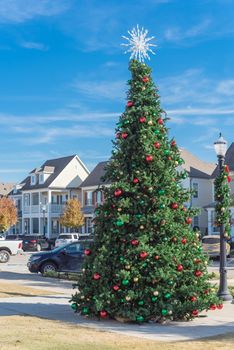 Giant Christmas tree with snowflake tree topper and colorful glass ornaments ball on display at City Square park in Coppell, Texas, USA. Xmas decoration with parked cars and country-style houses