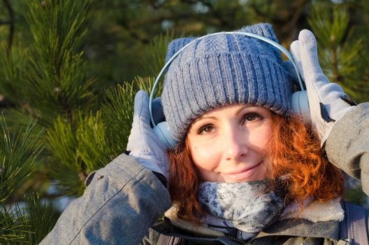 red-haired young girl in winter clothes listens to music with headphones on the background of pines