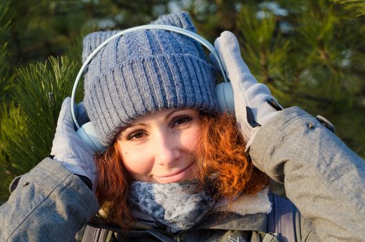 red-haired young girl in winter clothes listens to music with headphones on the background of pines