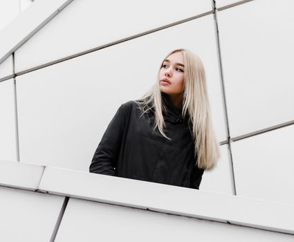 young blonde girl in black clothes against a gray building