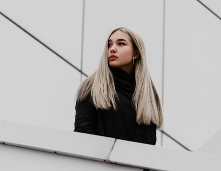 young blonde girl in black clothes against a gray building
