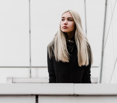 young blonde girl in black clothes against a gray building