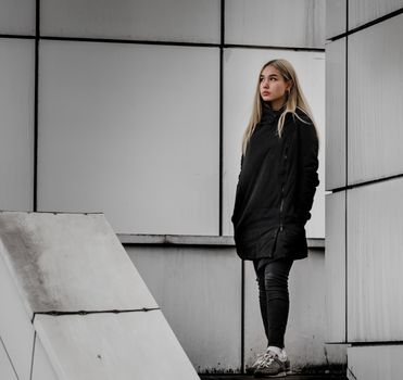 young blonde girl in black clothes against a gray building