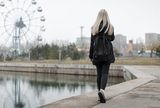 young girl with blond hair in black clothes and gray sneakers with a backpack goes along the river bank to an amusement park