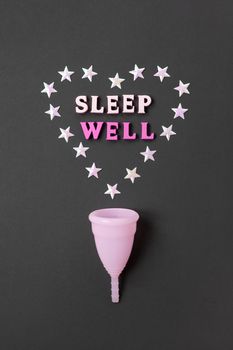 Pink menstrual cup on black background with with heart and words Sleep Well. Concept possibility of use cup at night, zero waste, savings, minimalism. Feminine hygiene product, flat lay. Vertical.